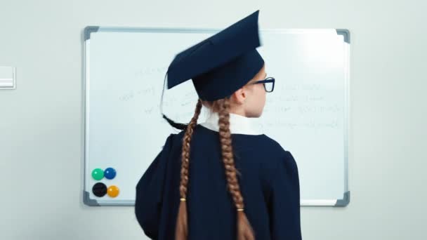 Cute student 7-8 years graduate in the mantle turns at camera holding book and smiling with teeth near whiteboard. Thumb up. Ok. Slider — Stock video