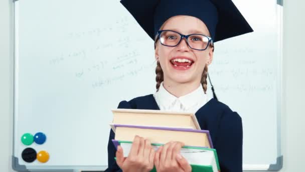 Estudiante de retrato de cerca 7-8 años se gradúa en el manto y sombrero sosteniendo libros y riéndose con dientes cerca de la pizarra. Pulgar hacia arriba. Bien, control deslizante. — Vídeo de stock