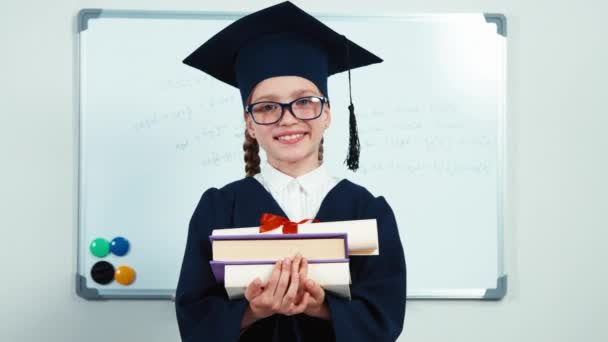 Primer plano retrato niña estudiante 7-8 años se gradúan en el manto sosteniendo sus libros y diploma y sonriendo con los dientes a la cámara — Vídeo de stock