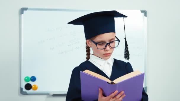 Gros plan portrait heureuse étudiante fille 7-8 ans diplômé dans le manteau et chapeau lire son livre et sourire à la caméra avec des dents — Video