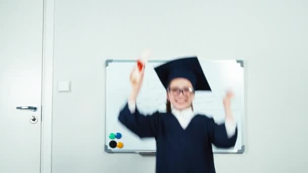 Graduate rejoices. Little student girl in the mantle holding her diploma and smiling at camera. Hands up — Stock video