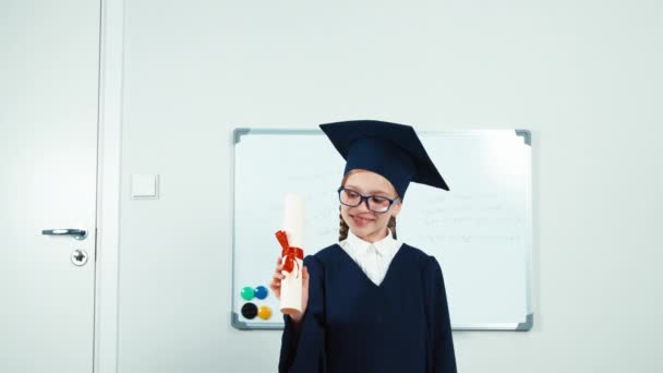 Pequeña estudiante graduada en el manto sosteniendo su diploma y sonriendo a la cámara. Manos arriba y riendo — Vídeo de stock