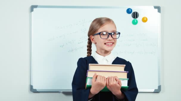 Retrato pequeña estudiante sosteniendo sus libros y sonriendo a la cámara con los dientes. Niña en gafas y manto de pie cerca de pizarra en el aula — Vídeo de stock