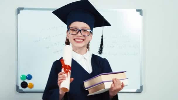 Close-up portrait little student girl 7-8 years graduate in the mantle holding her books and looking through diploma at camera with smile — Stock Video