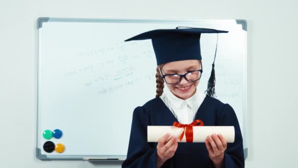 Close-up retrato menina estudante 7-8 anos de pós-graduação no manto beijando diploma e sorrindo com os dentes na câmera — Vídeo de Stock