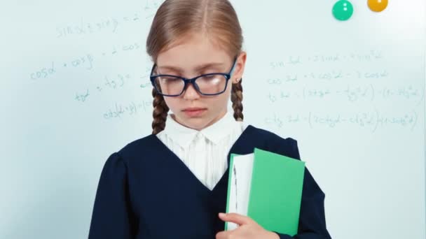 Gros plan portrait petite étudiante tenant son livre. Enfant fille en manteau debout près du tableau blanc dans la salle de classe et regardant la caméra — Video