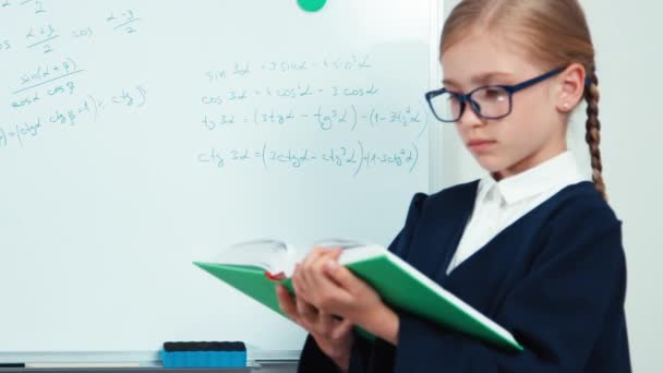 Portrait little student girl in glasses holding book standing near whiteboard and smiling at camera — Stock video