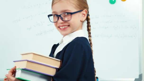 Close up portrait little student turns at camera and smiling with teeth. Child girl in glasses and mantle standing near whiteboard and holding books — ストック動画