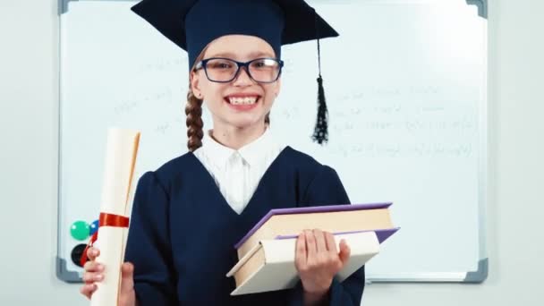 Primer plano retrato feliz estudiante chica 7-8 años se gradúan en el manto sosteniendo sus libros y mirando a través de diploma en la cámara con sonrisa — Vídeos de Stock