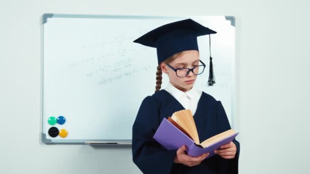 Graduate in the mantle hugging book. Close up portrait little student girl 7-8 years smiling at camera — Stock Video