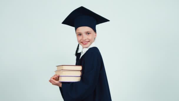 Graduate student girl 7-8 years in the mantle and hat turns around at camera with books on white and smiling at camera with teeth — Stock video