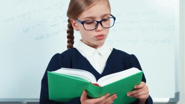 Nahaufnahme Porträt kleiner Schüler Lesebuch. Mädchen in Mantel steht neben Whiteboard im Klassenzimmer und lächelt mit Zähnen in die Kamera — Stockvideo