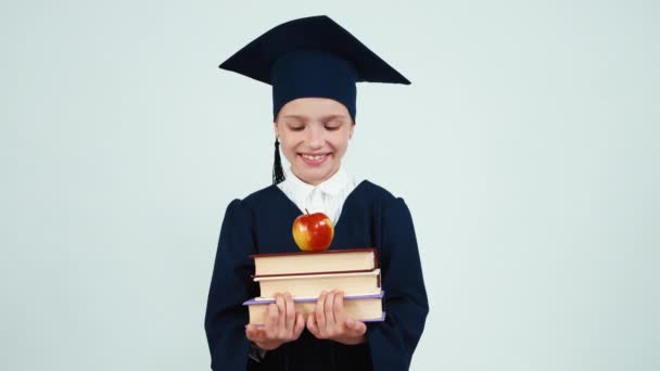 Primer plano retrato graduado chica 7-8 años en el manto y sombrero sosteniendo libros y manzana en blanco y sonriendo a la cámara con los dientes — Vídeos de Stock