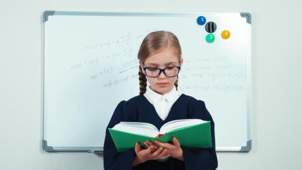 Close-up portret weinig student houden van haar boek. Kind meisje in mantel permanent in de buurt van whiteboard in de klas en lachend op camera — Stockvideo