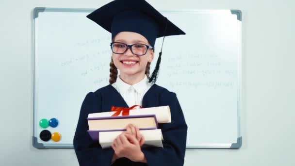 Gros plan portrait petite étudiante de 7-8 ans diplômée dans le manteau tenant ses livres et son diplôme et souriant avec des dents à la caméra. Pouce en l'air. — Video
