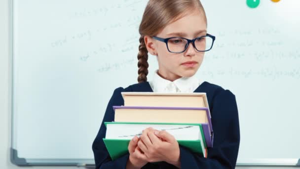 Chiudere ritratto piccolo studente sorridente con i denti alla macchina fotografica. Bambino ragazza in occhiali e mantello in piedi vicino alla lavagna bianca e libri in mano — Video Stock