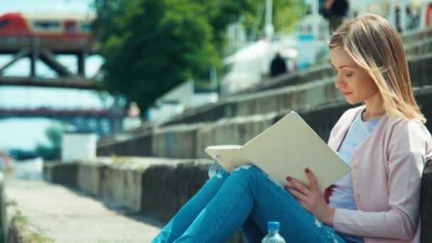 Giovane donna leggendo il tuo libro e seduto sulla banchina della città e guardando con sorriso alla macchina fotografica — Video Stock