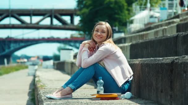 Mujer joven sentada en el muelle de la ciudad y mirando con sonrisa a la cámara — Vídeos de Stock