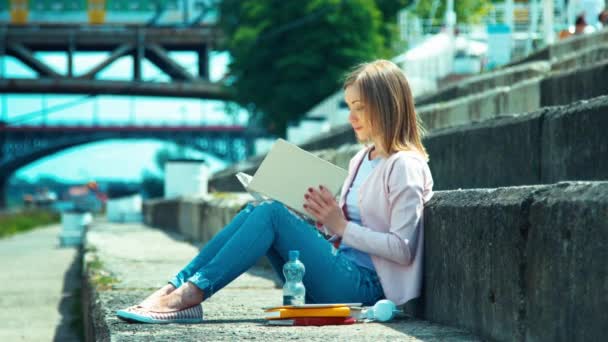 Молодая женщина открывает вашу книгу и сидит на городской набережной и смотрит с улыбкой в камеру — стоковое видео