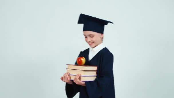 Primer plano retrato chica graduada 7-8 años en el manto y sombrero sosteniendo libros y manzana en blanco y sonriendo a la cámara con dientes. Niño entrando en el marco — Vídeo de stock