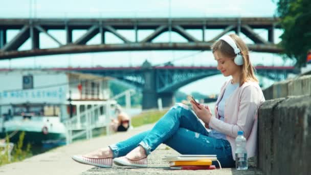 Young adult woman in headphones using mobile phone outdoors on city quay against bridge — Stock Video