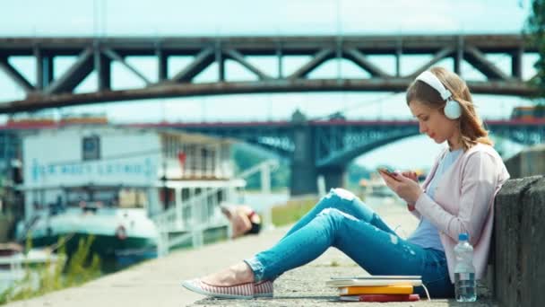 Giovane donna adulta utilizzando cuffie e telefono cellulare all'aperto sulla banchina della città contro il ponte — Video Stock