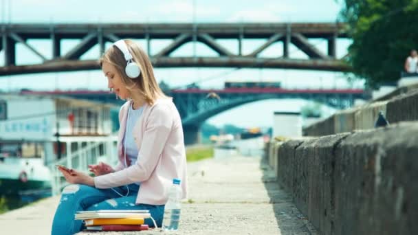 Νεαρή ενήλικη γυναίκα στα ακουστικά χρησιμοποιώντας το κινητό τηλέφωνο σε εξωτερικούς χώρους στην πόλη της αποβάθρας και τραγουδώντας το τραγούδι — Αρχείο Βίντεο
