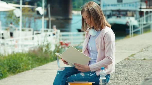 Ung vuxen kvinna med hörlurar läser bok utomhus ler mot kameran — Stockvideo