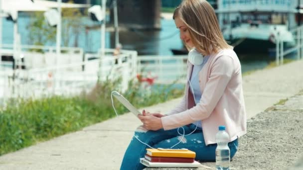 Młoda kobieta dorosłych ze słuchawkami za pomocą komputera typu tablet na zewnątrz w pobliżu rzeki. Uśmiecha się do kamery — Wideo stockowe