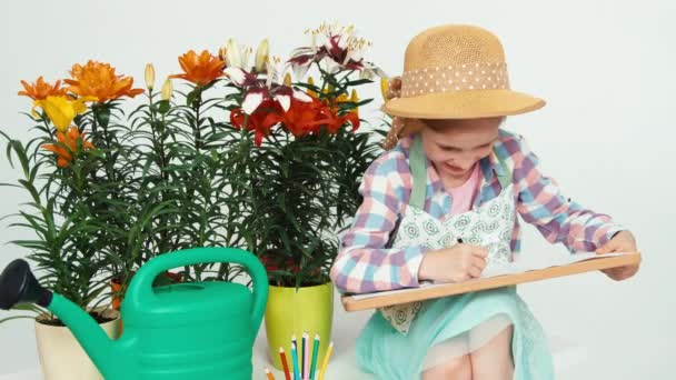 Ritratto fiore-ragazza bambino seduto vicino ai fiori e disegno su tavola da disegno e sorridente con i denti — Video Stock