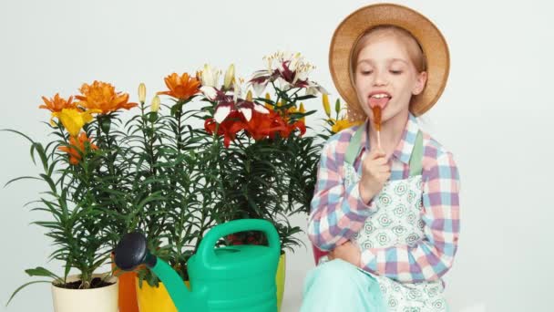 Närbild porträtt Blomsterflicka barnet sitter nära blommor och vattenkanna, hålla lollipop och ler mot kameran — Stockvideo