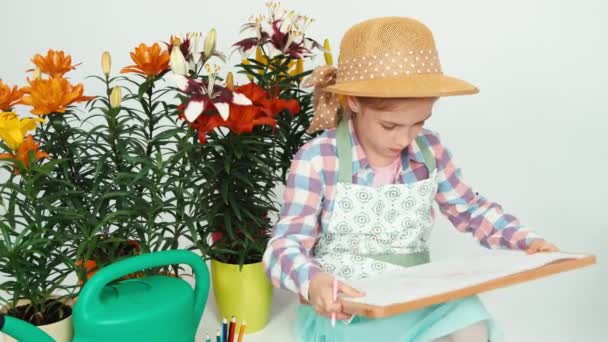 Крупним планом портрет дівчинки сидить біля квітів і малює на дошці для малювання і посміхається — стокове відео