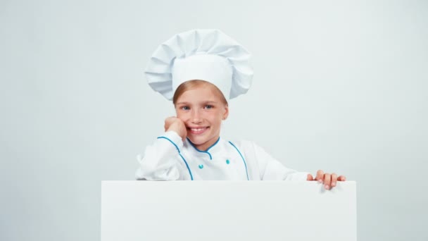 Chef chica en uniforme 7-8 años detrás de la pizarra blanca y tiene la mano cerca de la cara sonriendo a la cámara. Aislado sobre blanco — Vídeo de stock