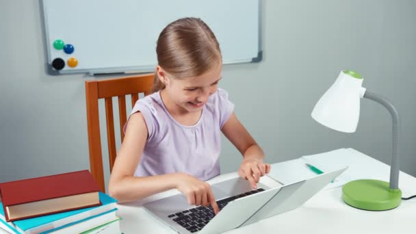 Menina 7-8 anos algo digitando no laptop sentado em sua mesa e sorrindo para a câmera. Vista superior — Vídeo de Stock