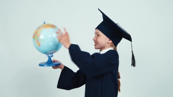 Close-up portret afgestudeerde meisje 7 tot 8 jaar in de mantel en hat holding globe op wit en lachen om de camera. Duim omhoog. OK — Stockvideo