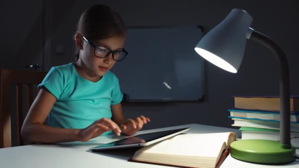Close up retrato escolar niña 7-8 años de edad en gafas usando tableta en la noche en su escritorio y sonriendo a la cámara — Vídeo de stock