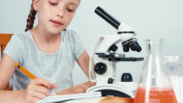 顕微鏡と彼女の演習本に書いて何かを使用して肖像画の学校の女の子を閉じます。白で隔離。パン — ストック動画