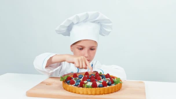 Close-up portret jonge baker onderzoekt en ontroerend vinger chocolade taart met snoep en fruit. Geïsoleerd op wit — Stockvideo