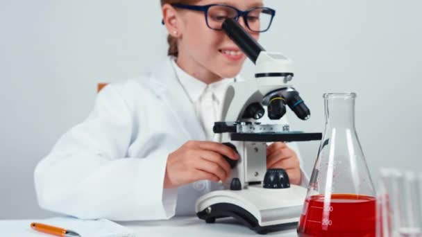 Gelukkig jonge chemicus school meisje 7-8 jaar met behulp van de Microscoop en schrijven in haar notitieblok en camera kijken. Geïsoleerd op wit — Stockvideo