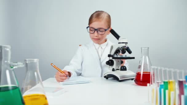 Счастливая школьница-химик 7-8 лет пользуется микроскопом, пишет в блокноте и улыбается в камеру. Изолированные на белом — стоковое видео