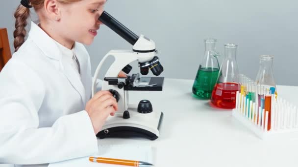 Retrato químico escolar niña 7-8 años utilizando microscopio y escritura en su cuaderno y sonriendo a la cámara con los dientes. Panorama — Vídeos de Stock