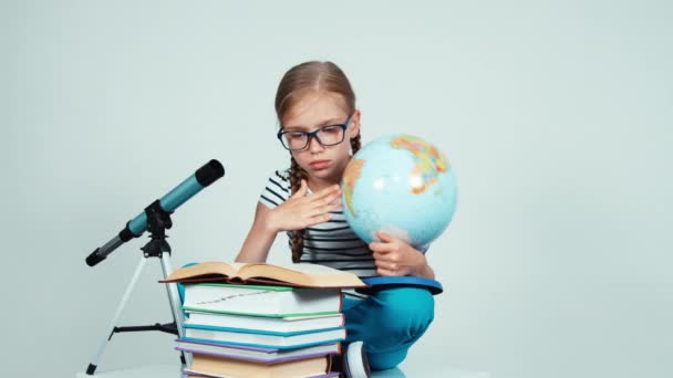 Portrait fille 7-8 ans livre de lecture et tenant son globe scolaire. Écolière en lunettes sur fond blanc assis sur le sol — Video