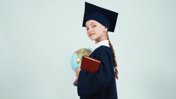 Портрет выпускницы девушка 7-8 лет в мантии и шляпе холдинг глобус и книги на белом фоне и улыбаясь на камеру с зубами — стоковое видео