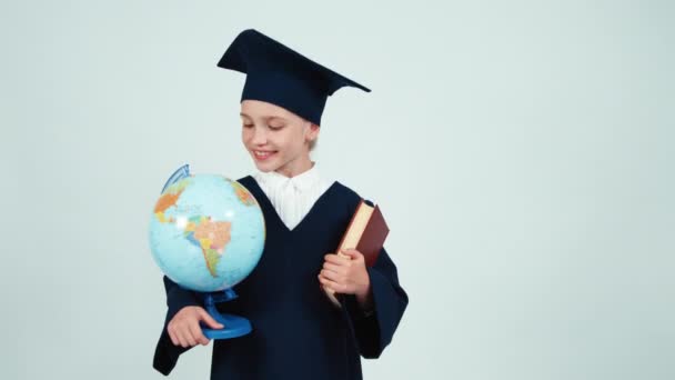 Ritratto laureata ragazza 7-8 anni nel mantello e cappello tenendo globo e libro su sfondo bianco e sorridente alla macchina fotografica — Video Stock