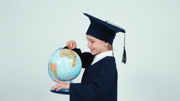 Portrait graduate girl 7-8 years in the mantle holding globe on white and smiling at camera — Stock Video