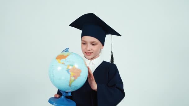 Portrait graduate girl 7-8 years in the mantle holds rotating globe on white and smiling at camera. Thumbs up. Ok — Stock Video