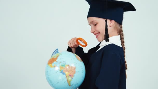 Portrait graduate girl 7-8 years in the mantle looking through magnifying glass globe on white background — Stock Video