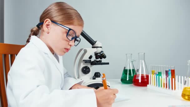Retrato en perfil joven niña de la escuela de química 7-8 años usando microscopio y escritura en su cuaderno. Aislado sobre blanco — Vídeos de Stock