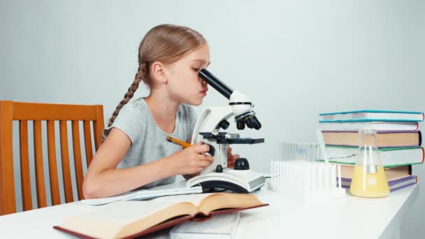 Портрет школьницы 7-8 лет с помощью микроскопа и что-то писать в ее тетрадь упражнений и улыбаясь на камеру — стоковое видео