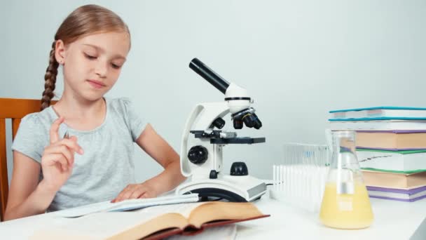 Chica de la escuela de retratos usando microscopio y algo escrito en su libro de ejercicios. Aislado sobre blanco — Vídeo de stock